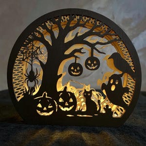 May include: A black, wooden Halloween-themed decoration in a semi-circle shape. The design features a tree, pumpkins, a spider, a cat, a ghost, and a raven. The interior is illuminated with warm light, creating a spooky ambiance.