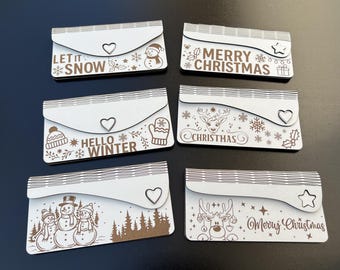 May include: A collection of six white wooden Christmas-themed gift card holders. Each holder features a unique design with winter and holiday-related text and illustrations, including snowmen, snowflakes, and reindeer.