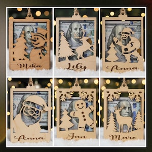Peut inclure: Ornements de Noël en bois avec un design découpé, chacun présentant un billet de 100 dollars visible à travers le cadre. Les motifs incluent un bonhomme de neige, un renne, le Père Noël, un bonhomme en pain d'épices et un cerf. Des noms sont gravés en bas.