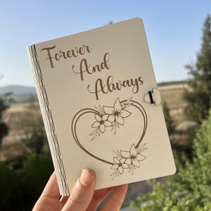 Op de afbeelding: Een lichtgekleurd houten tijdschrift met de woorden "Forever And Always" gegraveerd op de omslag. Een hartvormig bloemenontwerp is onder de tekst geëtst. Het tijdschrift heeft een sluiting.