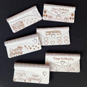 Może przedstawiać: Kolekcja drewnianych uchwytów na kartki okolicznościowe wyciętych laserowo z różnymi uroczystymi wiadomościami. Projekty obejmują "Happy Birthday", "Gratulacje z okazji ślubu" i "Happy Wedding Day" z elementami dekoracyjnymi.
