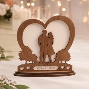 May include: A wooden heart-shaped photo frame with a silhouette of a couple embracing. The frame features a base with cut-out hearts and the names "JULIA & JACOB" engraved on a small plaque. Two trees flank the couple's silhouette.