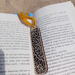 May include: A wooden bookmark with a detailed, intricate design cut out of the wood. The bookmark is light brown and has a yellow ribbon attached to the top.