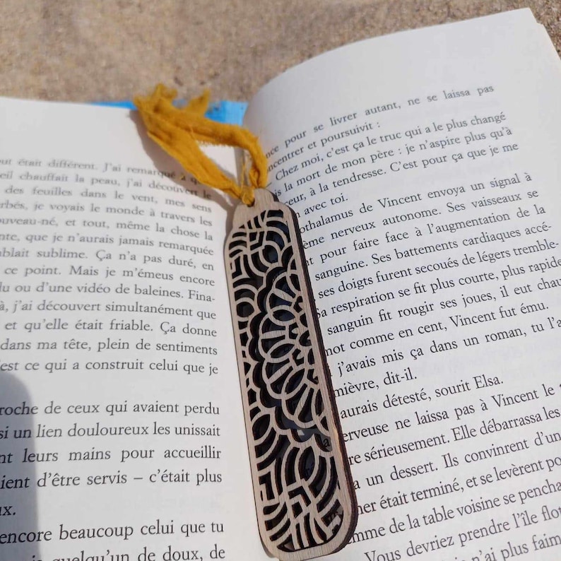 May include: A wooden bookmark with a detailed, intricate design cut out of the wood. The bookmark is light brown and has a yellow ribbon attached to the top.