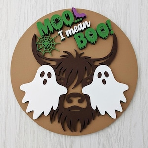 May include: A round, brown Halloween decoration with a brown Highland cow design and two white ghost figures. The text "Moo, I mean Boo!" is in green and white. A green spiderweb and purple bat are also included.