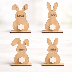 Peut inclure: Quatre décorations de Pâques en forme de lapin en bois. Chaque lapin est brun clair avec une queue blanche et duveteuse et un nom gravé sur le corps. Les noms sont Lisa, Jack, Sam et Mila. Chaque lapin repose sur une petite base rectangulaire.