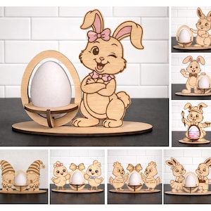 Pode incluir: Suporte de ovo de Páscoa de madeira com um coelhinho piscando com um laço rosa e um ovo decorativo. O suporte é feito de madeira clara e foi projetado para conter um ovo de tamanho padrão. Outras decorações de Páscoa de madeira também são mostradas.