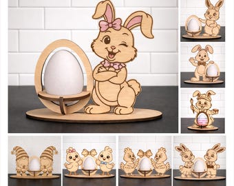 May include: Wooden Easter egg holder featuring a winking bunny with a pink bow and a decorative egg. The holder is made of light-colored wood and is designed to hold a standard-sized egg. Other Easter-themed wooden decorations are also shown.