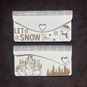 Op de afbeelding: Twee witte en bruine decoratieve enveloppen met winterthema's. Eén heeft de tekst "LET IT SNOW" en een sneeuwpop. De andere toont drie sneeuwpoppen en een bos silhouet. Elk heeft een hartvormige uitsparing.