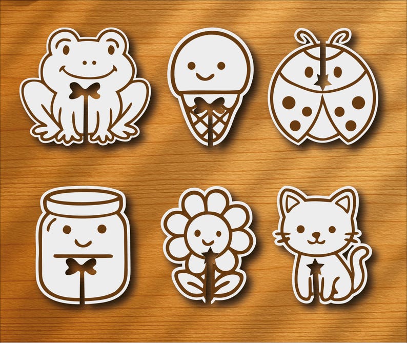 Op de afbeelding: Zes witte en bruine cartoonstickers op een houten oppervlak. De stickers bevatten een kikker, een ijsje, een lieveheersbeestje, een pot, een bloem en een kat. Elke sticker heeft een lachend gezicht en een bruine omtrek.