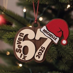 Könnte beinhalten: Weihnachtsbaumschmuck aus Holz mit dem Text "Seven IX 2025" und einem Weihnachtsmannhut mit der Aufschrift "Michelle". Der Schmuck ist braun, beige und rot und hängt an einer roten Schnur.