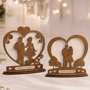 May include: Two wooden heart-shaped wedding decorations. Each features a silhouette of a couple inside the heart. One has the names "Karine & Enniko" and the other "Julia & Jacob". The decorations are brown with a white background.