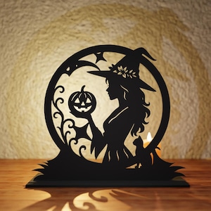 May include: Black silhouette of a witch holding a carved pumpkin, set within a circular frame with stars and decorative elements. A cat sits nearby. The design is a Halloween-themed decoration, possibly a candle holder.