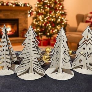 Puede incluir: Conjunto de cinco adornos navideños de madera con forma de árbol. Cada árbol está cortado con láser con diseños intrincados y se apoya sobre una base redonda. Un árbol tiene una estrella en la parte superior. Luces decorativas.