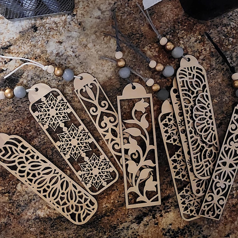 May include: A set of eight wooden bookmarks with intricate laser-cut designs. The bookmarks feature various patterns, including floral, geometric, and mandala designs. The bookmarks are made of light brown wood and have a natural finish.