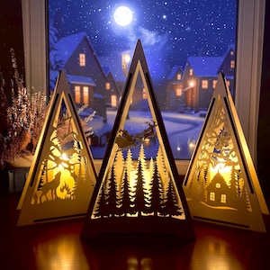 Puede incluir: Tres faroles de madera con forma de árbol de Navidad con intrincados diseños recortados. Cada farol presenta una escena invernal diferente, incluyendo un lobo, el trineo de Papá Noel y una casa. Los faroles están iluminados desde dentro, creando un brillo cálido.