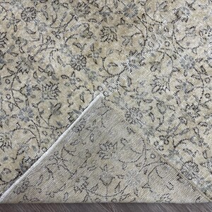 May include: A beige and gray floral patterned rug with a folded corner. The rug features a detailed design of flowers and vines in shades of gray and blue against a beige background. The rug's texture is visible.