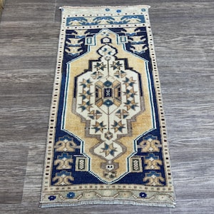 May include: A rectangular, hand-knotted rug with a central design in beige, navy blue, and cream. The rug features a geometric pattern with floral and star motifs, framed by a navy blue border. The rug is approximately 30 inches long.