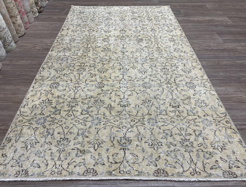 May include: A rectangular, vintage-style rug with an intricate floral pattern in shades of beige, gray, and blue. The rug has a distressed appearance, suggesting it is an antique or a reproduction with a worn look. The design features stylized flowers and vines.