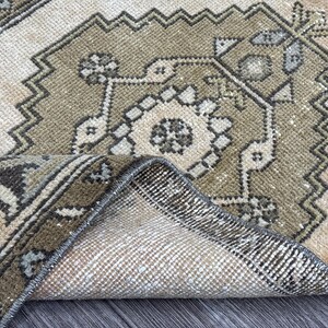 May include: A rolled-up, vintage-style rug with a detailed geometric pattern. The rug features a central design in shades of beige and olive green, framed by a darker border. The intricate design includes floral and angular shapes.
