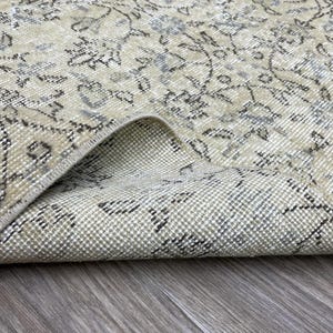 May include: Close-up of a beige and gray vintage rug with a floral pattern. The rug is folded over, revealing the underside. The rug's design features intricate floral motifs and a distressed appearance, suggesting an antique or vintage style.
