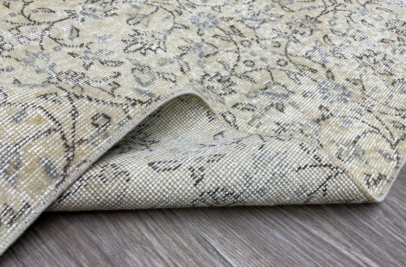 May include: Close-up of a beige and gray vintage rug with a floral pattern. The rug is folded over, revealing the underside. The rug's design features intricate floral motifs and a distressed appearance, suggesting an antique or vintage style.