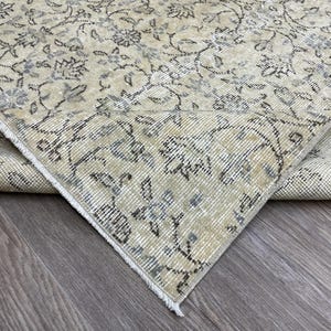May include: A beige and gray vintage rug with an intricate floral pattern. The rug's design features delicate flowers and vines in shades of gray and blue against a neutral background. The corner of the rug is folded over, revealing the underside.