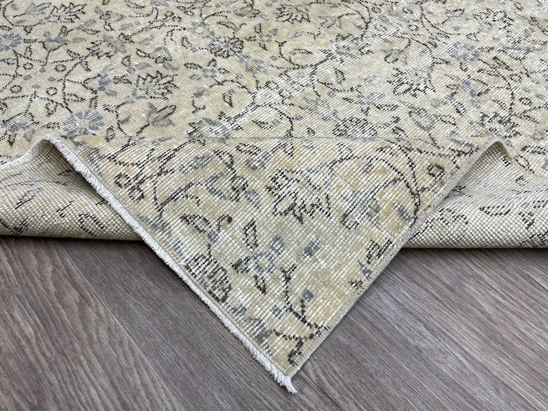 May include: A beige and gray vintage rug with an intricate floral pattern. The rug's design features delicate flowers and vines in shades of gray and blue against a neutral background. The corner of the rug is folded over, revealing the underside.
