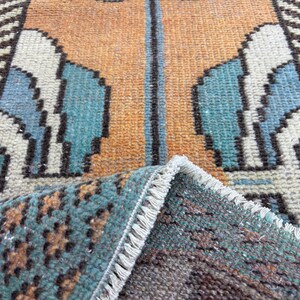 May include: Close-up of a vintage rug with a geometric pattern. The rug features a central design in shades of orange and teal, with white and brown accents. The edges are fringed, and a portion of the rug is folded over, revealing the underside.