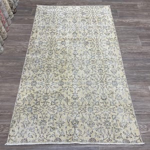 May include: Rectangular beige rug with an intricate floral pattern in shades of gray and blue. The rug is displayed on a dark wood floor, showcasing its detailed design and texture. Rolled rugs are visible in the background.