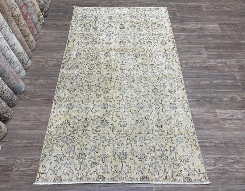 May include: Rectangular beige rug with an intricate floral pattern in shades of gray and blue. The rug is displayed on a dark wood floor, showcasing its detailed design and texture. Rolled rugs are visible in the background.