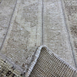 Puede incluir: Primer plano de una alfombra beige y marrón con un patrón tejido texturizado. La alfombra tiene un diseño de patchwork con rayas verticales y costuras visibles. La esquina de la alfombra está doblada, revelando la parte inferior.