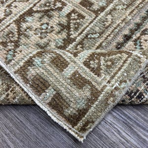 May include: Close-up of a folded, rectangular rug with a geometric pattern. The rug features a mix of brown, beige, and light blue colors. The edges are frayed, and the rug is resting on a wooden surface.