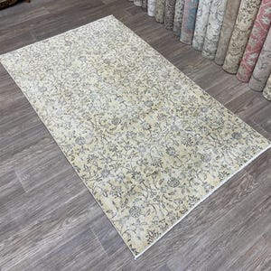 May include: Rectangular beige rug with an intricate floral pattern in shades of gray and blue. The rug is placed on a wooden floor, with rolled-up rugs visible in the background. The rug's design features a detailed, all-over floral motif.