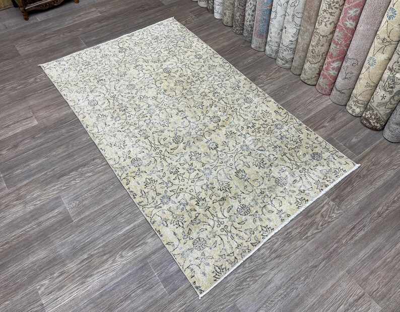 May include: Rectangular beige rug with an intricate floral pattern in shades of gray and blue. The rug is placed on a wooden floor, with rolled-up rugs visible in the background. The rug's design features a detailed, all-over floral motif.