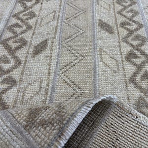 Puede incluir: Primer plano de una alfombra beige y marrón estampada. La alfombra presenta rayas verticales con diseños geométricos y florales estilizados en tonos marrones y beige. La textura de la alfombra es visible, con una esquina doblada que revela la parte inferior.