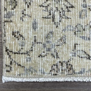 May include: Close-up of a section of a beige and gray patterned rug with a distressed look. The rug has a fringe on the bottom edge and is placed on a wooden surface.