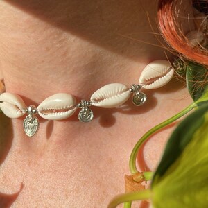 Può includere: Un girocollo di conchiglie bianche con ciondoli argentati. La collana è sul collo di una persona e lo sfondo è verde.