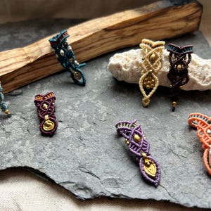 Könnte beinhalten: Eine Sammlung handgefertigter Makramee-Schmuckstücke in verschiedenen Farben, darunter Türkis, Lila, Orange und Gold. Die aufwendigen Designs weisen gewebte Muster und metallische Akzente auf, die auf einer grauen Steinoberfläche präsentiert werden.