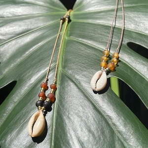 May include: Two necklaces with natural cowrie shell pendants and amber-colored beads. The necklaces are strung on thin, tan cords with gold-colored accents. The necklaces are displayed on a large, green leaf.