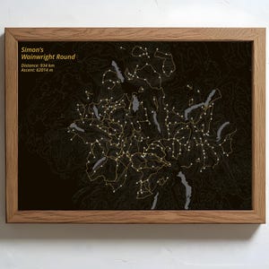 May include: Wooden framed print displaying a map of Simon's Wainwright Round. The map has a dark background with yellow lines and dots. Text at the top reads "Simon's Wainwright Round", with distance and ascent details.