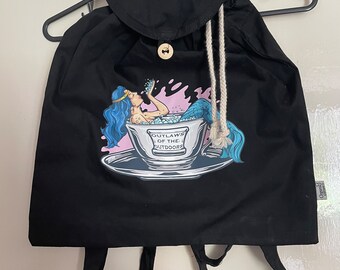 A black backpack bag with a mermaid having a bubble bath in a tea cup (outlaws of the outdoors) vinyl print on the front .