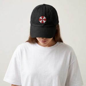 Puede incluir: Una gorra de béisbol negra con un logotipo bordado en forma de paraguas rojo y blanco centrado en el panel frontal. La gorra tiene una visera curva y una corona estructurada, confeccionada en tela negra sólida con costuras a juego en todo el diseño.