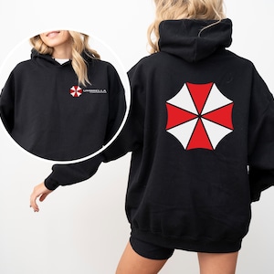 Puede incluir: Sudadera con capucha negra con un logotipo de paraguas rojo y blanco en la espalda y el texto "Umbrella Corporation" en la parte delantera.