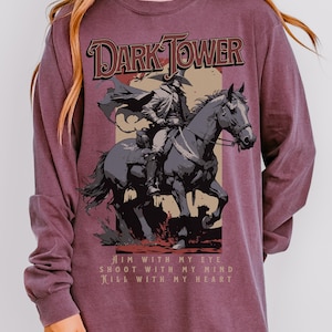 May include: A long-sleeved, maroon shirt featuring a graphic of a rider on horseback with the text "DARK TOWER" above and the quote "AIM WITH MY EYE SHOOT WITH MY MIND KILL WITH MY HEART" below. The design is in shades of gray and brown.