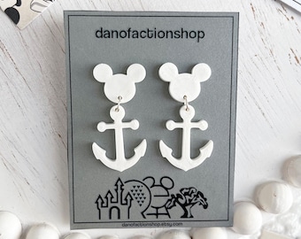 Puede incluir: Pendientes blancos colgantes con una silueta de cabeza de Mickey Mouse y un dije de ancla. Los pendientes se muestran en una tarjeta gris con el texto "danofactionshop" y una pequeña imagen de un castillo, una noria y un árbol.