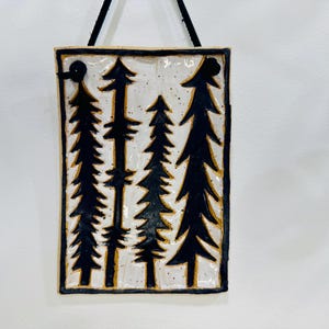 May include: Rectangular ceramic wall hanging depicting a forest scene with black fir trees on a white background. The trees are outlined in black, with a textured, speckled surface. The piece is framed with a dark border and has a black cord for hanging.