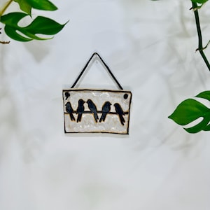 May include: A rectangular ceramic wall hanging with five black bird silhouettes on a line. The background is textured white with a black border and a black string for hanging. The piece is mounted on a white wall.