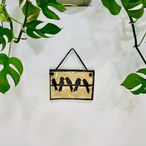 May include: A rectangular wall hanging with a black frame and a beige background. Five black bird silhouettes are perched on a black line. The sign is suspended by a black string. Green leafy plants frame the sign.