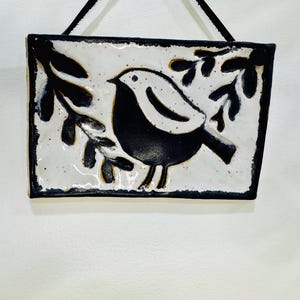 May include: A rectangular ceramic wall hanging with a black and white bird design. The bird is in the centre on a white background, with black leaves. The edges are black, and a black cord is attached for hanging.